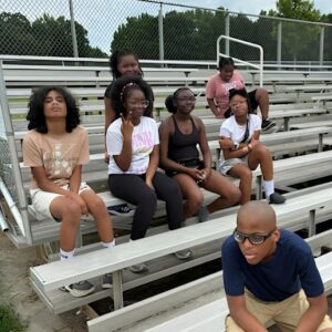 A group of students sits together on the bleachers during outdoor Olympics day games at Above and Beyond Students' free summer camp, Ranson Middle School, Charlotte, NC.
